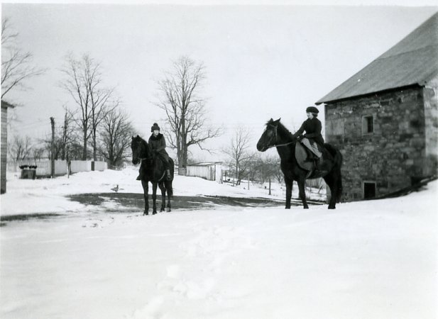 Photo of Ellen and Velva Henry on horseback with snow on the ground and a stone barn in the background