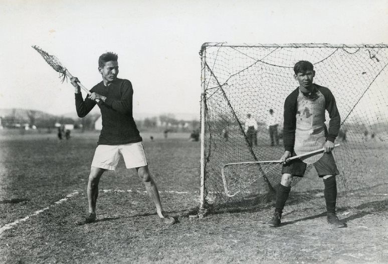 Photograph of  Sundown and Johns, lacrosse players, one demonstrating proper shooting technique while the other tends goal.